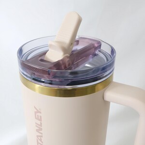 May include: A beige Stanley travel mug with a clear lid and a flip-top closure. The mug has a gold band around the middle and a handle. The word "STANLEY" is printed vertically on the side of the mug.