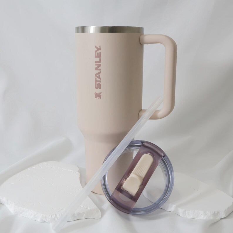 Pu&ograve; includere: Un bicchiere termico Stanley beige con manico, cannuccia e coperchio. Il bicchiere ha il logo Stanley stampato sul lato. Il coperchio &egrave; trasparente con un bordo viola e un cursore bianco. &Egrave; inclusa una cannuccia trasparente.