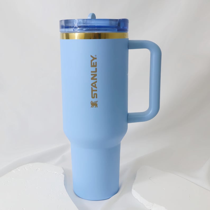 Op de afbeelding: Een lichtblauwe Stanley reismok met een handvat en een doorzichtig deksel. De mok heeft een gouden band aan de bovenkant en het Stanley-logo in goud. De mok is ontworpen om een grote hoeveelheid vloeistof te bevatten.