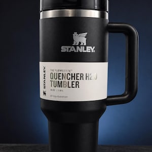 Może przedstawiać: Czarny kubek Stanley Quencher H2.0 z uchwytem. Kubek ma czarną pokrywkę i białą etykietę z napisem "QUENCHER H2.0 TUMBLER" i "1,18 L". Logo Stanley jest widoczne na kubku.