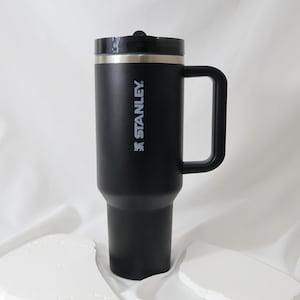 May include: Black Stanley insulated travel mug with a handle and a black lid. The mug has a silver rim and the Stanley logo printed in white. The mug is designed to keep beverages hot or cold for hours.