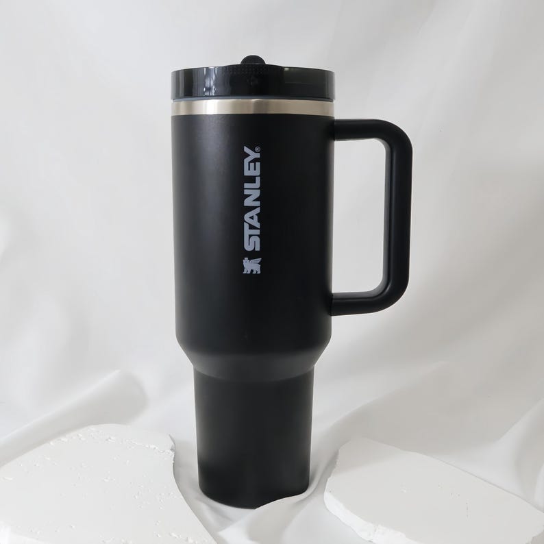 May include: Black Stanley insulated travel mug with a handle and a black lid. The mug has a silver rim and the Stanley logo printed in white. The mug is designed to keep beverages hot or cold for hours.