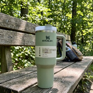 Op de afbeelding: Een groene Stanley Quencher H2.0 Tumbler met een wit deksel en handvat staat op een houten bank. De tumbler heeft het Stanley-logo en productinformatie erop gedrukt. De achtergrond bevat bomen en een rugzak.