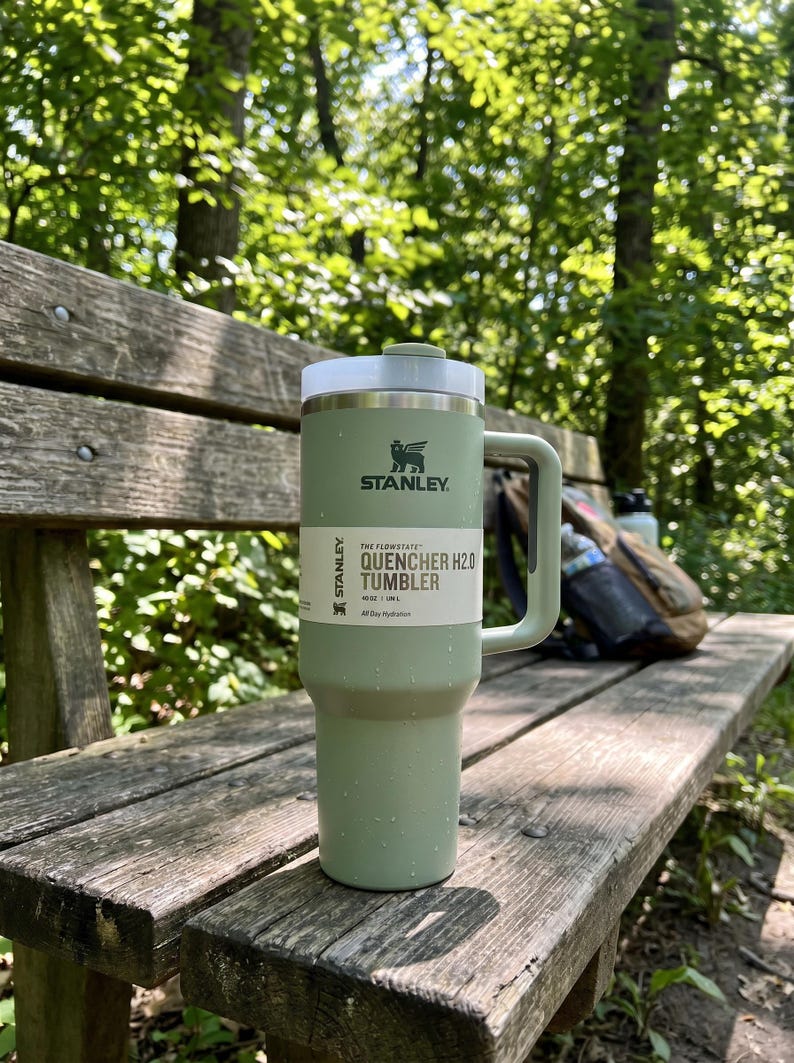 Op de afbeelding: Een groene Stanley Quencher H2.0 Tumbler met een wit deksel en handvat staat op een houten bank. De tumbler heeft het Stanley-logo en productinformatie erop gedrukt. De achtergrond bevat bomen en een rugzak.