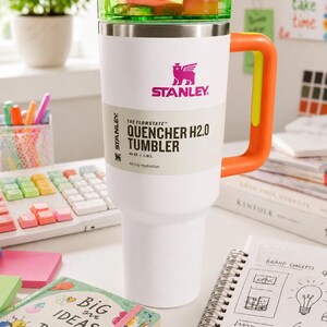 Pu&ograve; includere: Bicchiere Stanley Quencher H2.0 bianco con coperchio verde e rosa e manico arancione. Il bicchiere ha il logo Stanley e le informazioni sul prodotto stampate sopra. Il bicchiere &egrave; da 1,18 L.