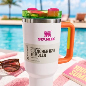 Pu&ograve; includere: Bicchiere Stanley Quencher H2.0 bianco con coperchio verde e rosa e manico arancione. Il bicchiere ha il logo Stanley e il testo "QUENCHER H2.0 TUMBLER" e "1,18 L". Il bicchiere &egrave; su un asciugamano a righe rosa e bianche.