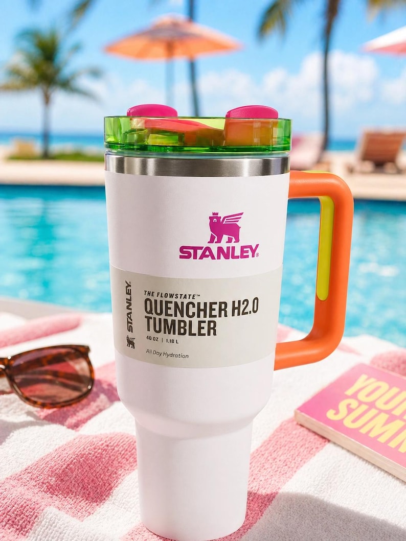 Pu&ograve; includere: Bicchiere Stanley Quencher H2.0 bianco con coperchio verde e rosa e manico arancione. Il bicchiere ha il logo Stanley e il testo "QUENCHER H2.0 TUMBLER" e "1,18 L". Il bicchiere &egrave; su un asciugamano a righe rosa e bianche.