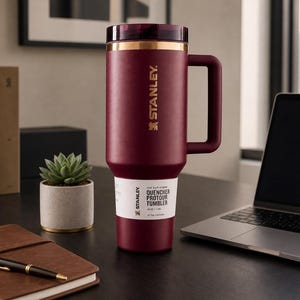 May include: A maroon Stanley Quencher Protour tumbler with a handle, gold accents, and the Stanley logo. The tumbler sits on a desk next to a laptop, a succulent plant, and a leather-bound notebook.
