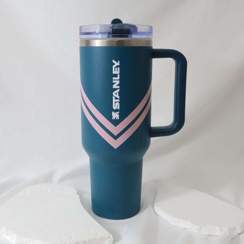 May include: A teal Stanley tumbler with a handle and a clear lid. The tumbler features a pink chevron design and the Stanley logo in white. The tumbler is designed to keep beverages cold or hot for hours.