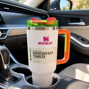 Pu&ograve; includere: Bicchiere Stanley Quencher H2.0 bianco con coperchio verde e rosso, manico arancione e logo Stanley. Il bicchiere &egrave; in un portabicchieri per auto. Il testo sul bicchiere recita "QUENCHER H2.0 TUMBLER" e "All Day Hydration".