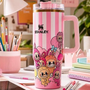 Pu&ograve; includere: Un bicchiere Stanley a righe rosa e bianche con manico, con un design a cartoni animati di personaggi mostruosi. Il bicchiere ha un coperchio e un manico rosa e il logo Stanley &egrave; visibile. Lo sfondo include cancelleria e una scrivania.
