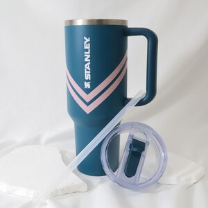 May include: A teal Stanley tumbler with a handle, a clear lid, and a straw. The tumbler has a silver rim and a pink and white chevron design. The Stanley logo is printed on the side.
