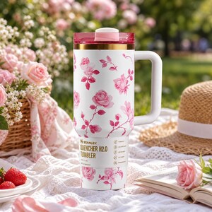May include: White Stanley Quencher H2.0 tumbler with a pink floral design. The tumbler has a handle, a pink lid, and gold accents. The tumbler is sitting on a white lace blanket with a picnic setting.