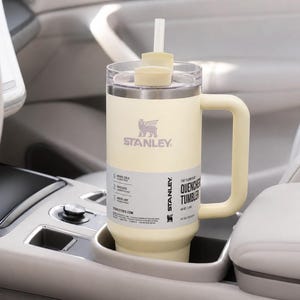 May include: A cream-colored Stanley Quencher tumbler with a handle and straw, resting in a car's cup holder. The tumbler features the Stanley logo and product information. The tumbler is designed to keep drinks cold for 2 days and iced for 4 days.