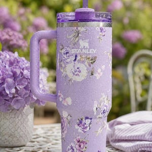 May include: A lavender Stanley tumbler with a floral design featuring white, purple, and pink flowers. The tumbler has a handle, a clear lid, and a purple straw. The Stanley logo is visible on the side.