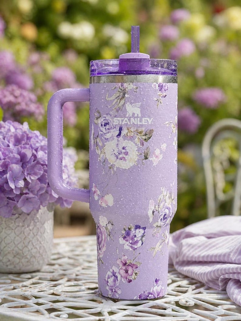 May include: A lavender Stanley tumbler with a floral design featuring white, purple, and pink flowers. The tumbler has a handle, a clear lid, and a purple straw. The Stanley logo is visible on the side.