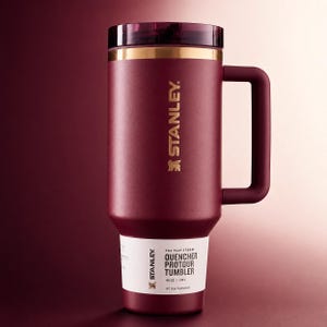 May include: A maroon Stanley Quencher Protour tumbler with a handle. The tumbler has a gold band near the top and a dark lid. The text "STANLEY" is printed in gold on the side. The bottom of the tumbler has a white label with product information.