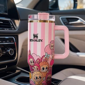 Pu&ograve; includere: Un bicchiere da viaggio Stanley a righe rosa e bianche con manico, con un'illustrazione di cartoni animati di creature stravaganti. Il bicchiere &egrave; nel portabicchieri di un'auto. Il logo Stanley &egrave; visibile.