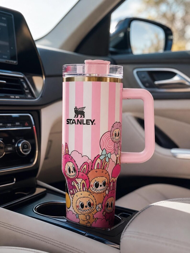 Pu&ograve; includere: Un bicchiere da viaggio Stanley a righe rosa e bianche con manico, con un'illustrazione di cartoni animati di creature stravaganti. Il bicchiere &egrave; nel portabicchieri di un'auto. Il logo Stanley &egrave; visibile.