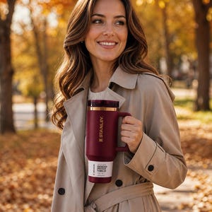 May include: A maroon Stanley Quencher ProTour tumbler with a gold rim and handle is held by a person wearing a tan trench coat. The tumbler has the Stanley logo and the text "QUENCHER PROTOUR TUMBLER" on a white label.