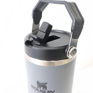 Op de afbeelding: Een grijze Stanley-beker met een zwart deksel en handvat. De beker heeft een flip-top opening en een zilveren rand. Het Stanley-logo is zichtbaar op de voorkant van de beker.
