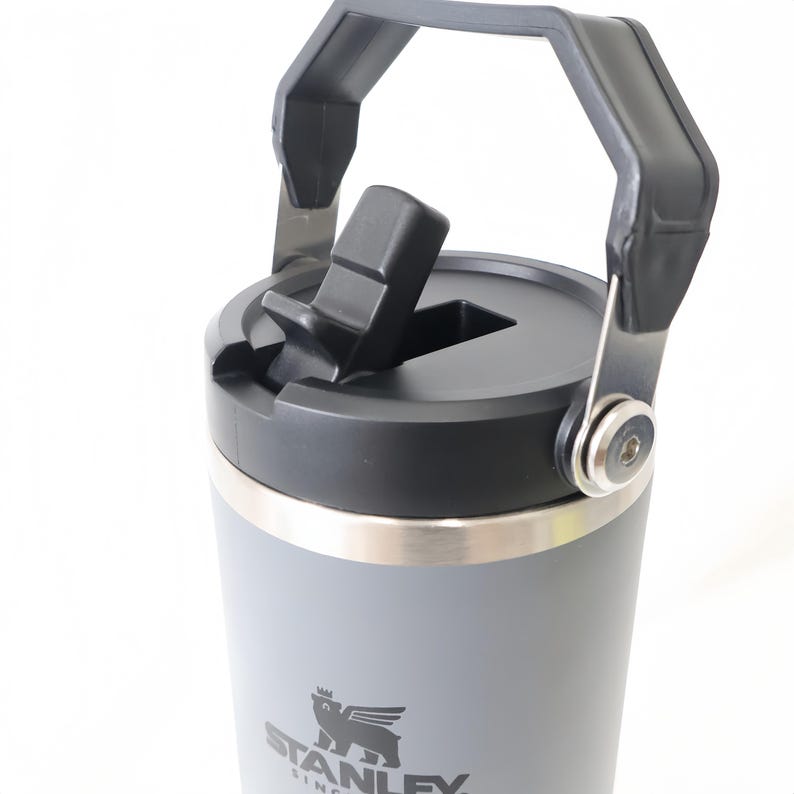Op de afbeelding: Een grijze Stanley-beker met een zwart deksel en handvat. De beker heeft een flip-top opening en een zilveren rand. Het Stanley-logo is zichtbaar op de voorkant van de beker.