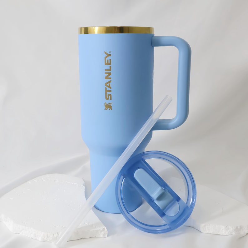 Op de afbeelding: Een lichtblauwe Stanley-beker met een gouden rand, een handvat en de merknaam erop. De beker heeft een blauw deksel en een doorzichtig rietje. Ontworpen voor koude dranken.