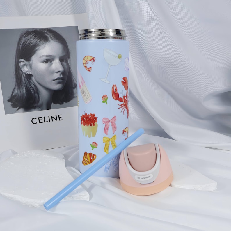 May include: A light blue insulated tumbler with a silver lid, decorated with illustrations of food and drinks, including a lobster and cocktail. A light blue straw and a pink and beige device are next to the tumbler.