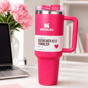 Puede incluir: Un vaso Stanley Quencher H2.0 de color rosa brillante con asa y tapa transparente. El vaso tiene el logotipo de Stanley y el texto "QUENCHER H2.0 TUMBLER" en una etiqueta blanca. Capacidad: 1,18L.