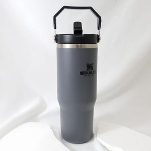 Op de afbeelding: Een grijze ge&iuml;soleerde beker met een zwarte handgreep en deksel. De beker is voorzien van het Stanley-logo en de tekst. De beker is ontworpen om dranken koud of warm te houden. De beker is ongeveer 25 cm hoog.