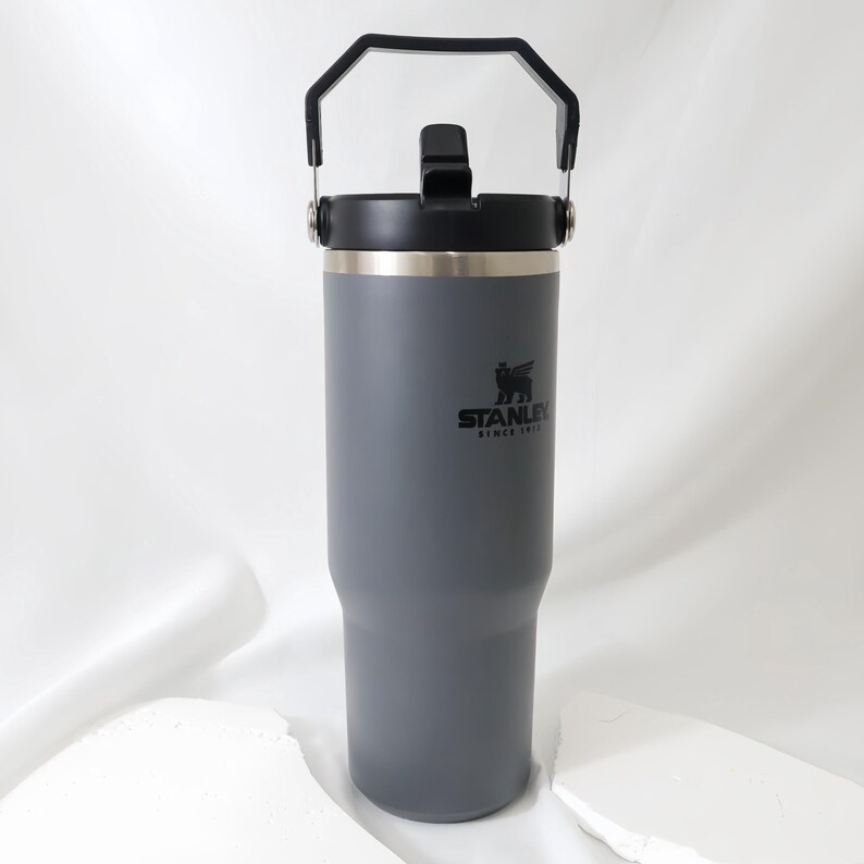 Op de afbeelding: Een grijze ge&iuml;soleerde beker met een zwarte handgreep en deksel. De beker is voorzien van het Stanley-logo en de tekst. De beker is ontworpen om dranken koud of warm te houden. De beker is ongeveer 25 cm hoog.