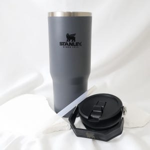 Op de afbeelding: Een grijze Stanley-beker met een zwart deksel, rietje en een zwarte handgreep. De beker heeft het Stanley-logo en de tekst "STANLEY. SINCE 1913" erop. Ontworpen om dranken koud of warm te houden. De beker is ongeveer 23 cm hoog.
