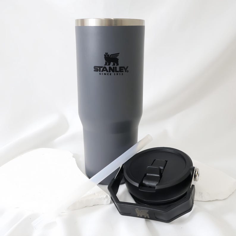 Op de afbeelding: Een grijze Stanley-beker met een zwart deksel, rietje en een zwarte handgreep. De beker heeft het Stanley-logo en de tekst "STANLEY. SINCE 1913" erop. Ontworpen om dranken koud of warm te houden. De beker is ongeveer 23 cm hoog.