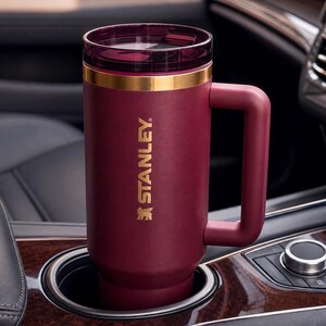 May include: A burgundy Stanley travel mug with a gold band and handle, resting in a car's cup holder. The mug has a clear lid and the Stanley logo in gold. The interior of the car is visible in the background.