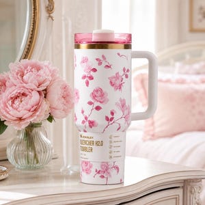 May include: White Stanley Quencher H2.0 tumbler with a pink floral pattern. The tumbler has a pink lid and handle, with gold accents. The design features pink roses and green leaves. The tumbler is on a white surface next to a vase of pink peonies.
