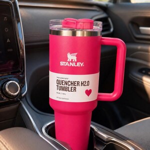 Puede incluir: Un vaso Stanley Quencher H2.0 rosa brillante, 1,18 L, con asa, est&aacute; en el portavasos de un coche. El vaso tiene una banda blanca con el texto "QUENCHER H2.0 TUMBLER" y un s&iacute;mbolo de coraz&oacute;n. El logotipo de Stanley es visible.