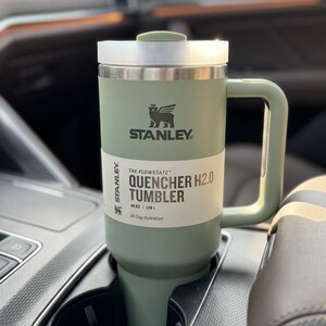Op de afbeelding: Een groene Stanley Quencher H2.0 tumbler, ongeveer 1,18 liter, met een witte en zilveren deksel en handvat, in een bekerhouder in de auto. De tumbler heeft het Stanley-logo en de tekst "All Day Hydration."