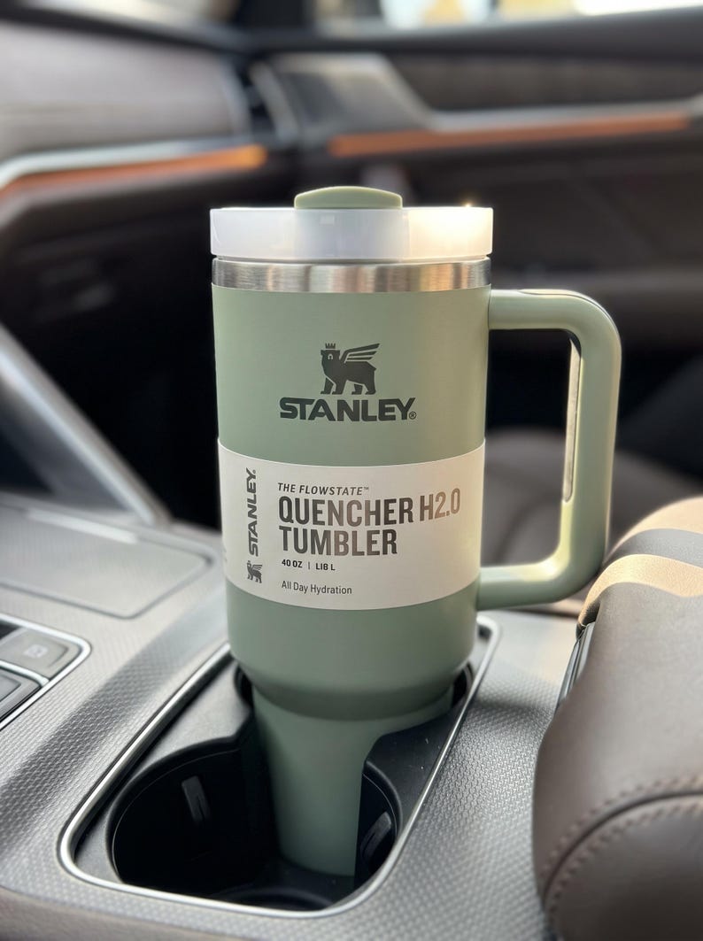Op de afbeelding: Een groene Stanley Quencher H2.0 tumbler, ongeveer 1,18 liter, met een witte en zilveren deksel en handvat, in een bekerhouder in de auto. De tumbler heeft het Stanley-logo en de tekst "All Day Hydration."