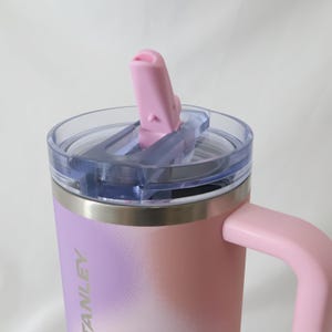 Op de afbeelding: Een roze en paarse ge&iuml;soleerde beker met een handvat en een doorzichtig deksel. Het deksel heeft een roze flip-top tuit. De beker heeft het woord "STANLEY" op de zijkant. Ontworpen om dranken warm of koud te houden.