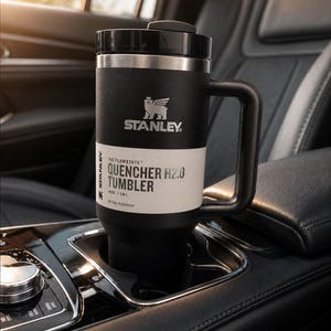 Może przedstawiać: Czarny kubek Stanley Quencher H2.0 z uchwytem, w uchwycie na kubek w samochodzie. Kubek ma czarną pokrywkę i biały pasek z napisem "QUENCHER H2.0 TUMBLER". Widoczne logo Stanley. Pojemność: 1,18 L.