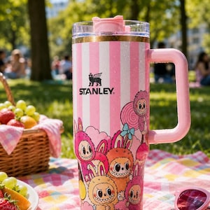 Pu&ograve; includere: Un bicchiere Stanley a righe rosa e bianche con manico, con un design a cartoni animati di mostri. Il bicchiere ha un coperchio trasparente e il logo Stanley. Lo sfondo mostra un picnic con frutta e occhiali da sole.