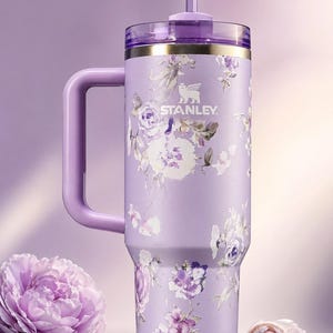 May include: A lavender Stanley tumbler with a floral design featuring white and purple flowers. The tumbler has a handle, a straw, and a clear lid with a gold rim. The Stanley logo is visible on the front.