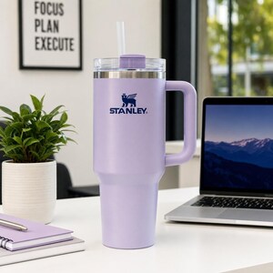 Puede incluir: Un vaso Stanley lavanda con asa y tapa transparente, con pajita. El logotipo de Stanley es visible en la parte delantera. El vaso est&aacute; sobre un escritorio blanco, con un port&aacute;til y una peque&ntilde;a planta en maceta en el fondo. El texto "FOCUS PLAN EXECUTE" est&aacute; en un marco.