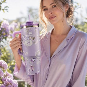 May include: A lavender Stanley tumbler with a floral design. The tumbler has a handle, a straw, and a purple lid. The floral pattern includes purple, white, and lavender flowers. The tumbler is held in front of a background of purple flowers.