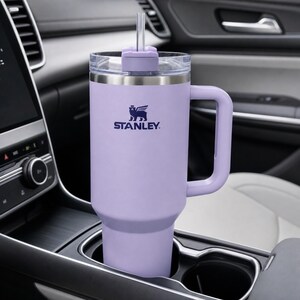 Puede incluir: Un vaso Stanley lavanda con asa y tapa transparente con pajita. El logotipo de Stanley est&aacute; impreso en azul marino. El vaso est&aacute; en el portavasos de un coche.