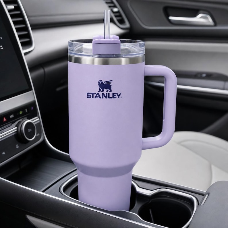 Puede incluir: Un vaso Stanley lavanda con asa y tapa transparente con pajita. El logotipo de Stanley est&aacute; impreso en azul marino. El vaso est&aacute; en el portavasos de un coche.