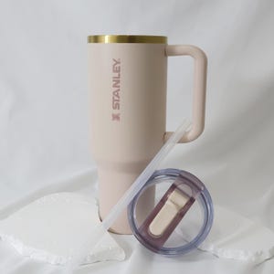 May include: A light pink Stanley tumbler with a gold rim and handle. The tumbler includes a clear straw and a lid with a light beige slider. The Stanley logo is printed on the side. The tumbler is approximately 9 inches tall.