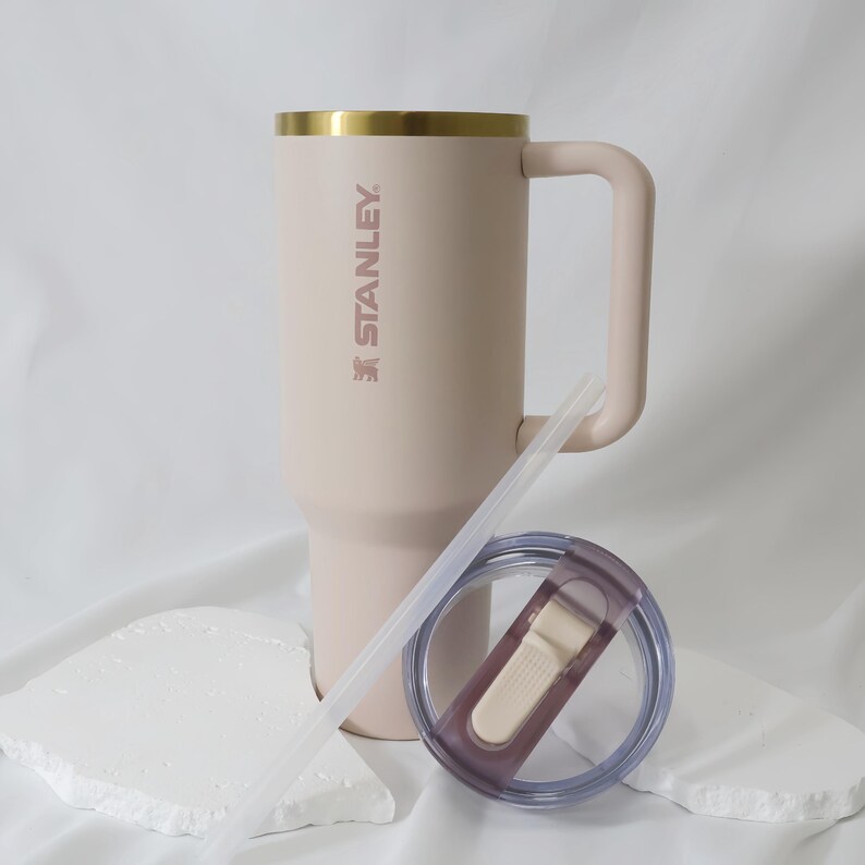 May include: A light pink Stanley tumbler with a gold rim and handle. The tumbler includes a clear straw and a lid with a light beige slider. The Stanley logo is printed on the side. The tumbler is approximately 9 inches tall.