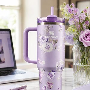 May include: A lavender Stanley tumbler with a floral design. The tumbler has a handle, a lid, and a straw. The Stanley logo is visible. The tumbler is on a desk with a laptop, pen, and a vase of flowers.
