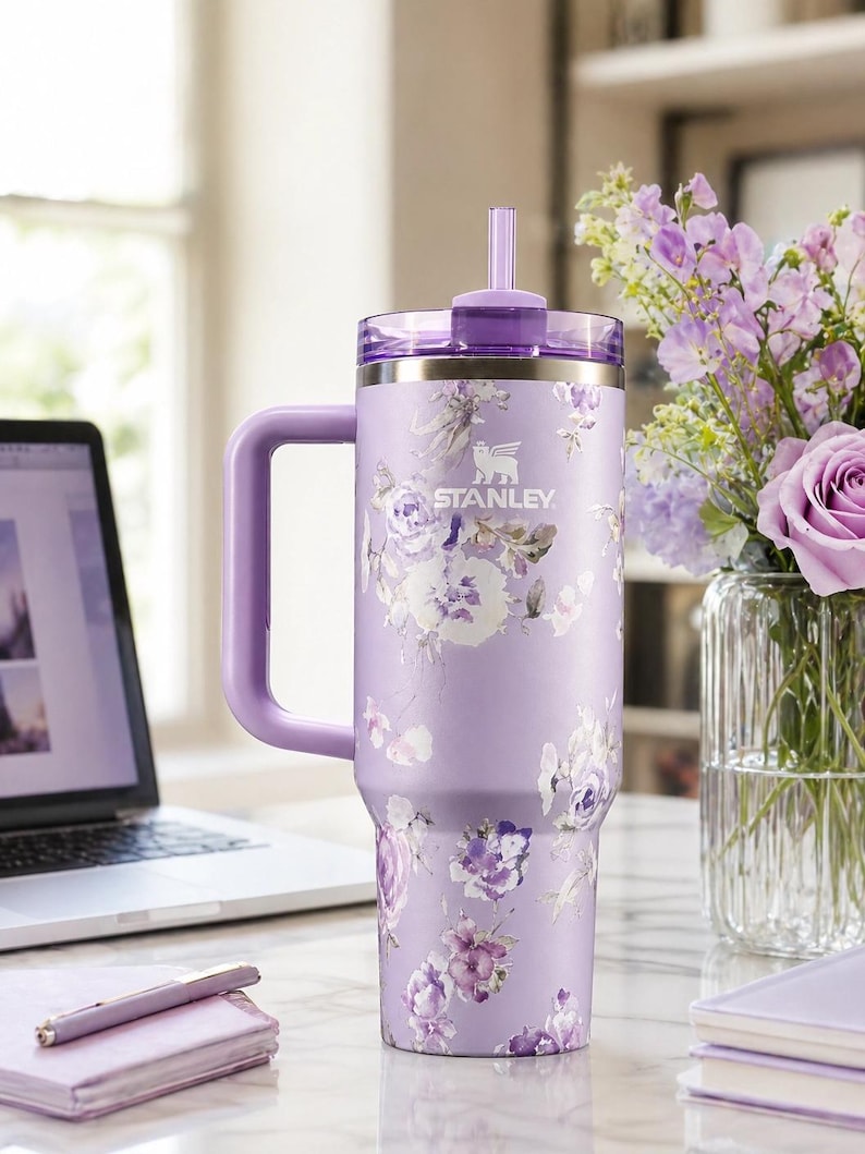 May include: A lavender Stanley tumbler with a floral design. The tumbler has a handle, a lid, and a straw. The Stanley logo is visible. The tumbler is on a desk with a laptop, pen, and a vase of flowers.